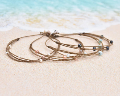 Tula Blue's waterproof Riptide anklet options in white pearls, blush pearls, larimar stones and peacock pearls on the sand by the surf.