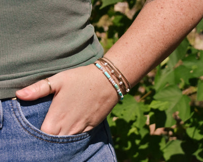 Turquoise, Pearl and Sterling Silver bracelet mixed with other bracelets on models wrist with her hand in her pocket in front of greenery